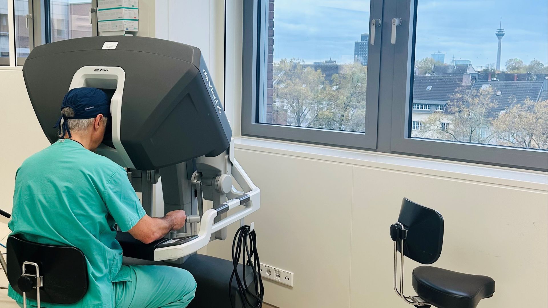 Der Chirurg sitzt im OP-Saal am Da Vinci OP-Roboter und bedient ihn währen einer Operation. Im Hintergrund sieht man durch die Fenster über die Stadt Düsseldorf.